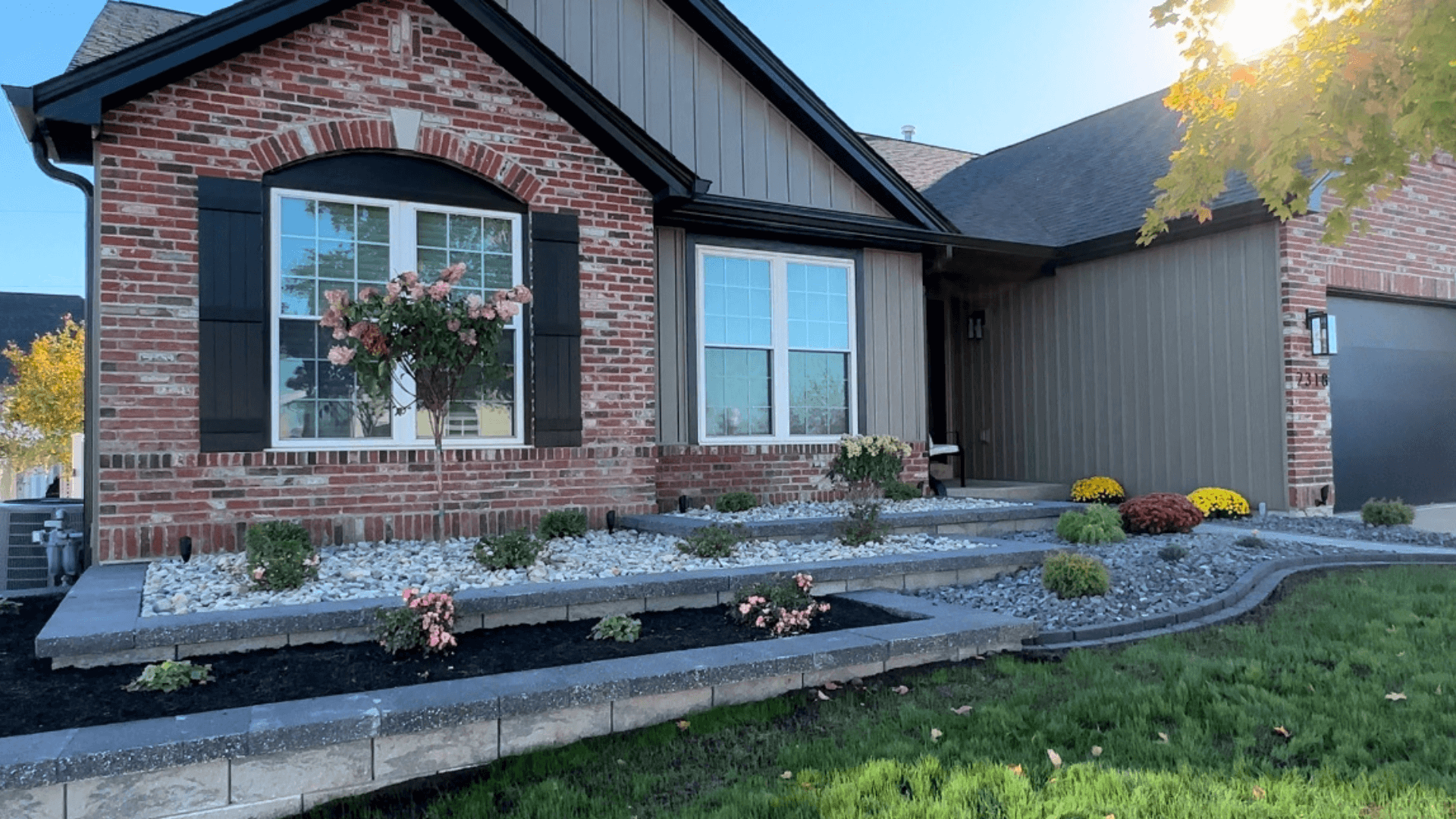 Front yard stone retaining wall raised planting bed with flowering shrubs and mulch at brick home by NxGen Outdoors