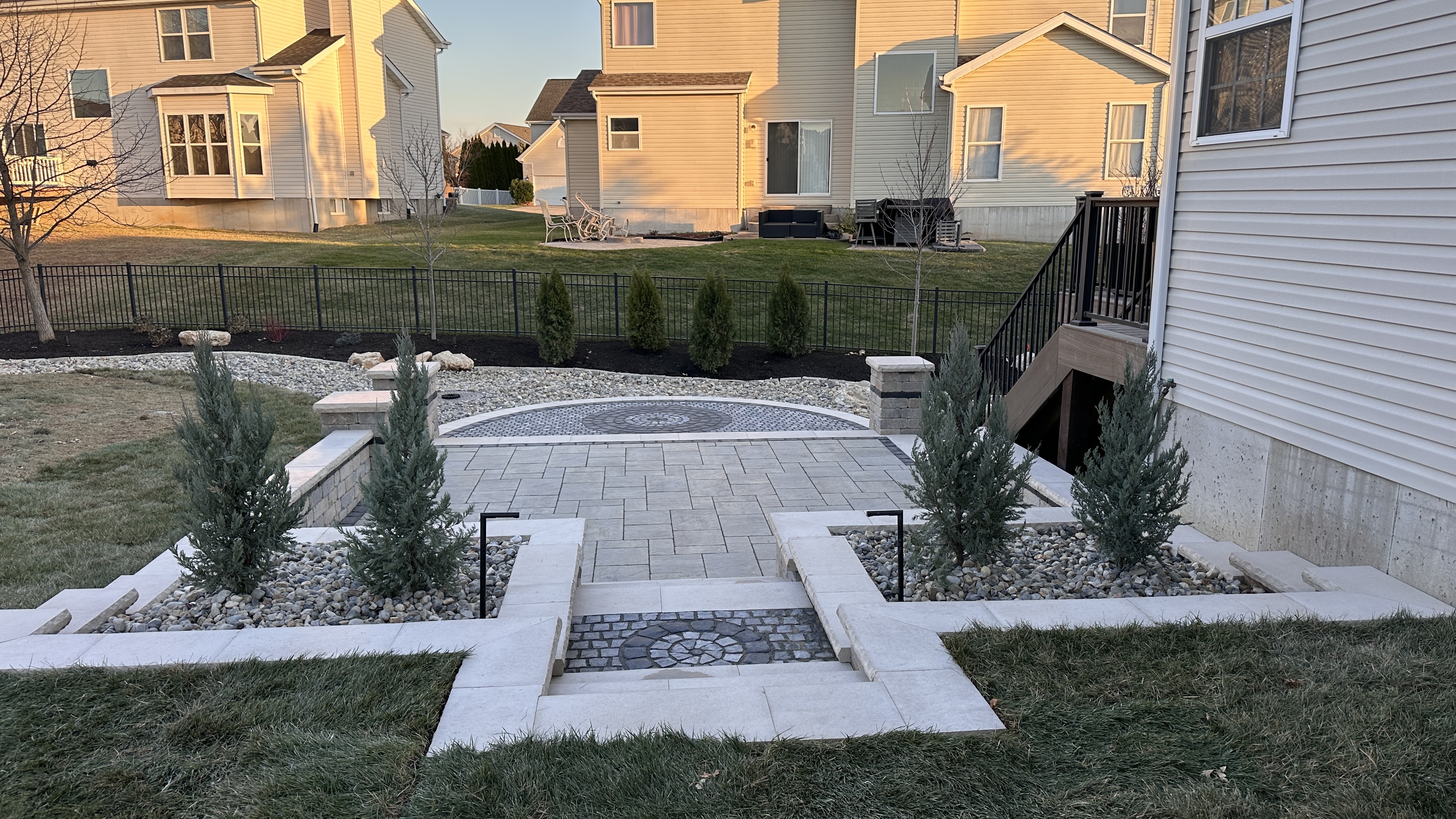 Wide-angle daytime view of completed paver patio with circular cobblestone inlay, stone seat walls, pillar, and evergreen landscaping by NxGen Outdoors
