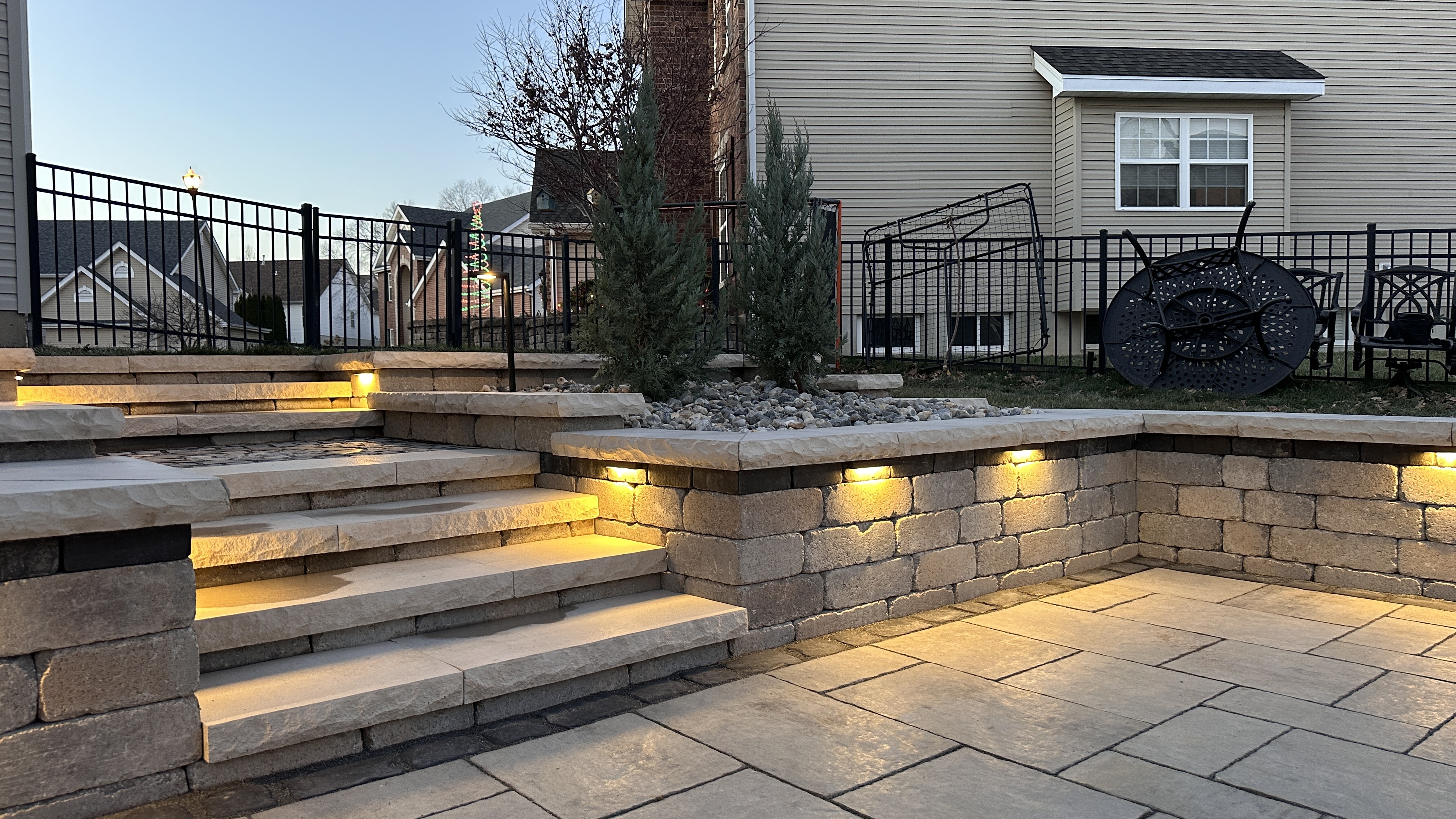 Looking up at LED-lit stone steps, seat walls with cap lights, and deck from the paver patio at night by NxGen Outdoors