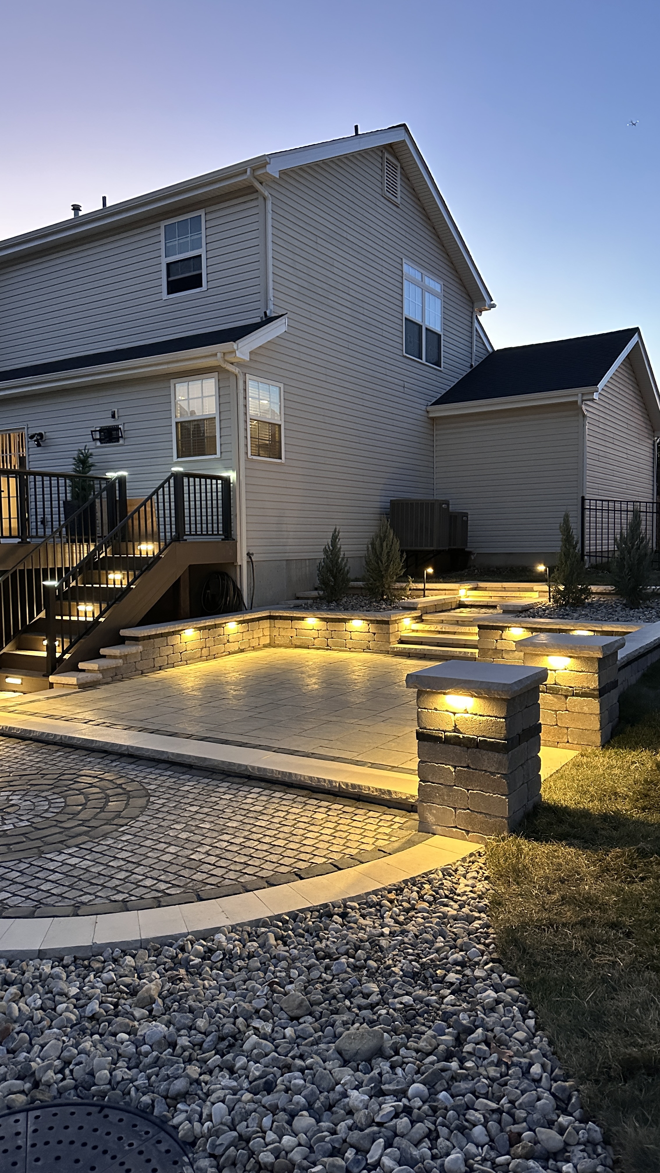 Nighttime view of stone steps with LED-lit column pillars and paver patio with landscape lighting by NxGen Outdoors